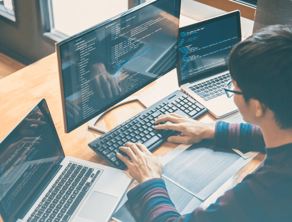 Developer coding at a desk with three screens and two laptops displaying source code; hands type on a keyboard in a focused, productive workspace.