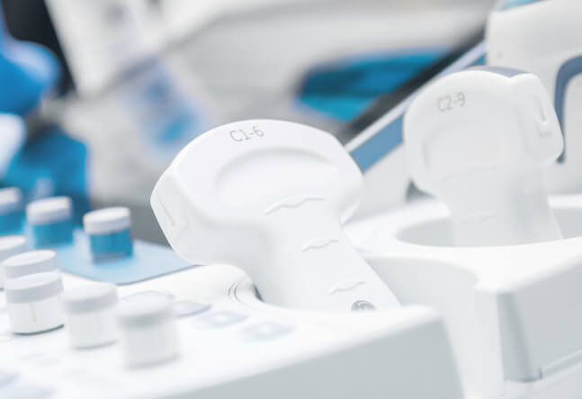 Close-up of ultrasound machine probes in a clinical setting. The white transducers are placed in their holders, conveying a clean, professional environment.