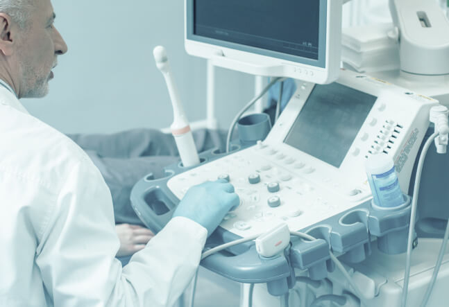 A medical professional in a white coat operates an ultrasound machine, pressing buttons and reviewing the monitor. The setting is clinical and focused.