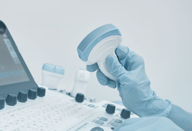 A gloved hand holds an ultrasound probe above a control panel, suggesting a medical setting. The tone is clinical and focused on healthcare technology.