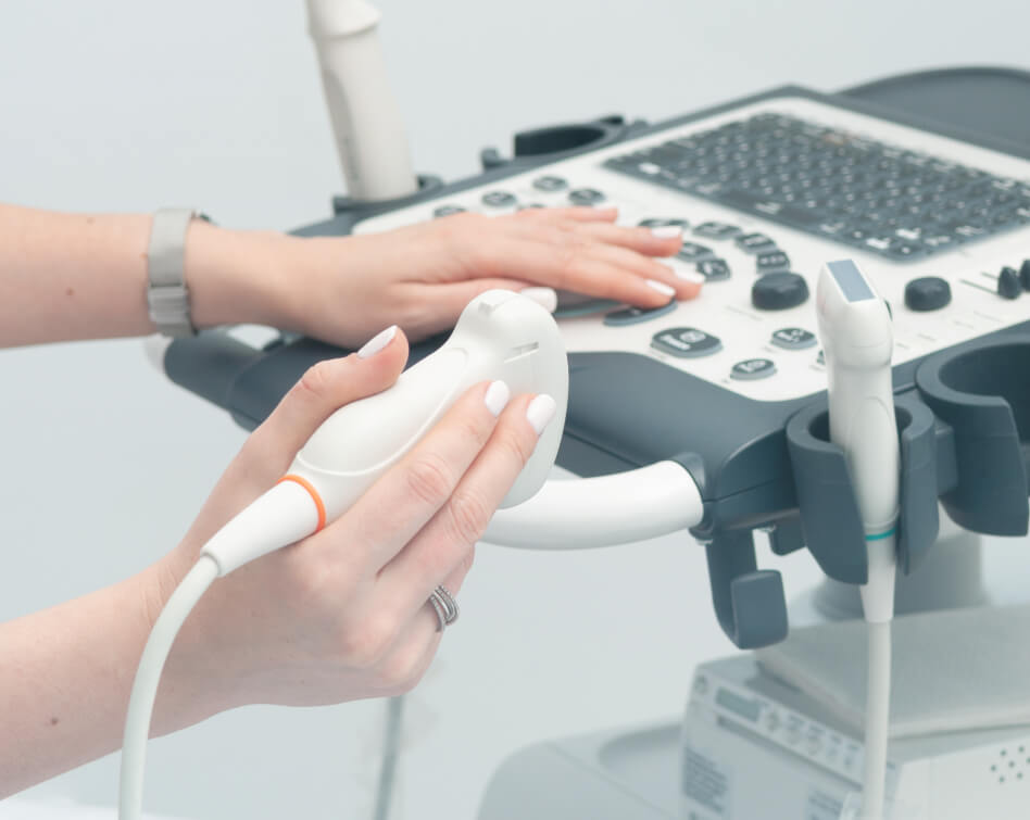 Hands adjust controls on an ultrasound machine, one holding a probe. The setting is clinical, suggesting a medical or diagnostic environment.
