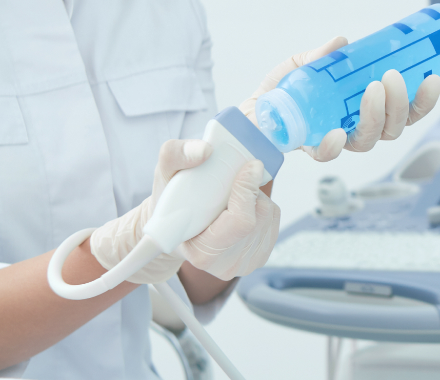 A healthcare professional applies gel from a blue bottle onto a medical ultrasound probe, wearing white gloves, indicating a clinical setting.