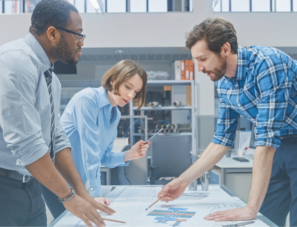 Three diverse coworkers collaborate over architectural plans in a bright office, pointing at charts and discussing a project with focused energy.