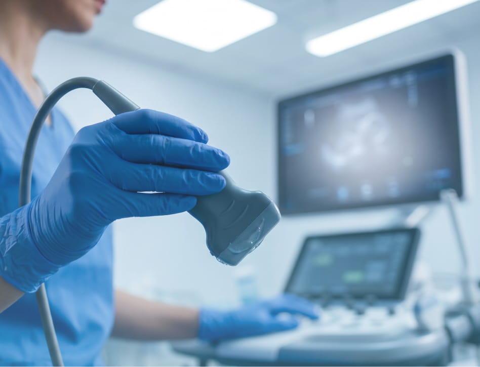 A medical professional in blue scrubs holding an ultrasound probe, with a monitor displaying sonogram images of a fetus.