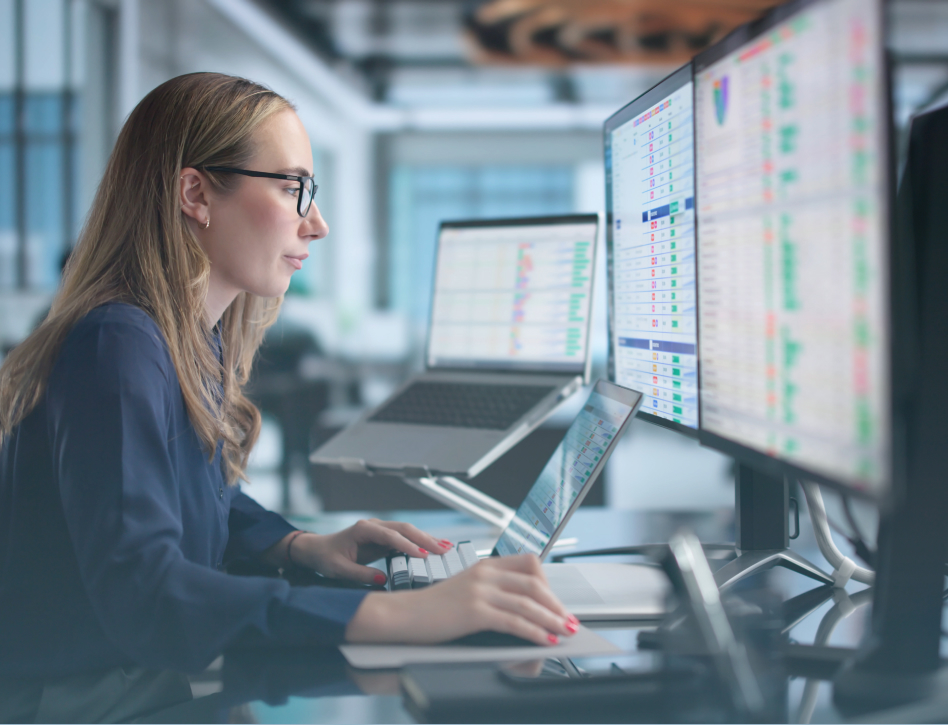 Focused woman in glasses works at a desk with multiple monitors displaying colorful charts and data in a modern office.