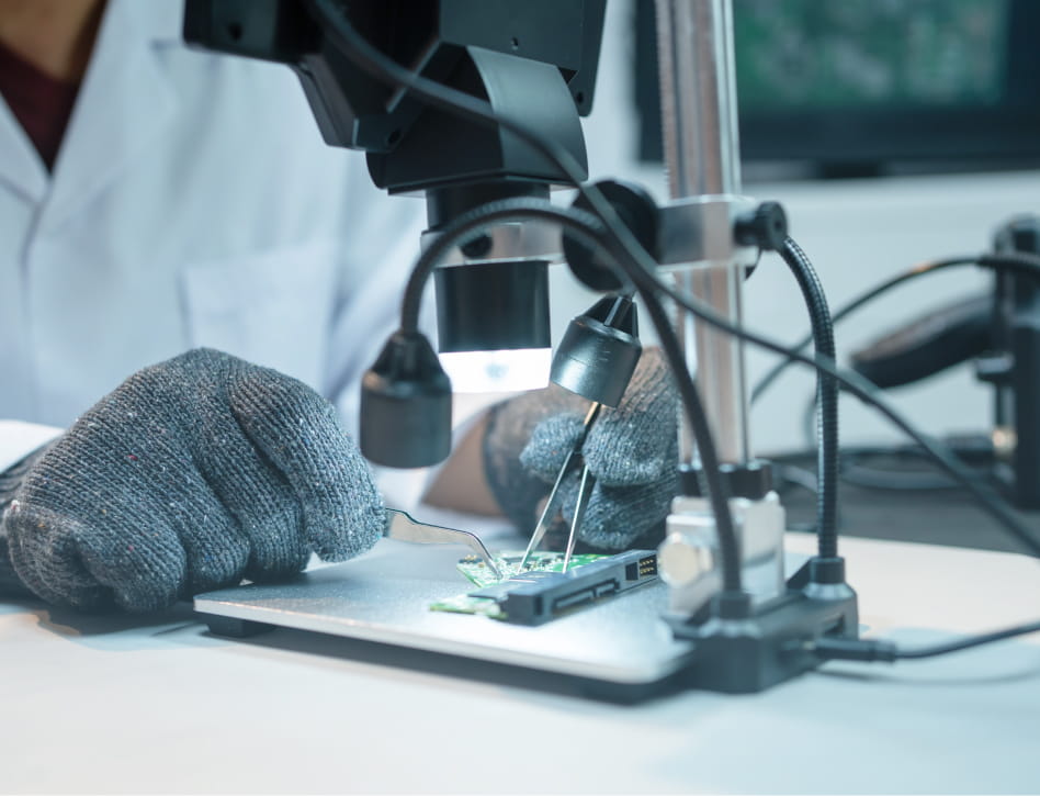 Close-up of a person in a lab coat using tweezers to work on a green circuit board under a microscope with LED lights.