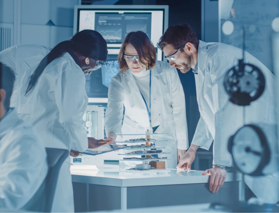 Three scientists in lab coats and safety glasses examine electronic components on a white table in a high-tech lab with data displays in the background.
