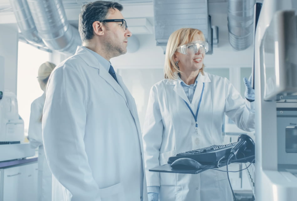 Two scientists in a modern laboratory, a man and a woman, observe a piece of equipment on the right as the woman points toward it.