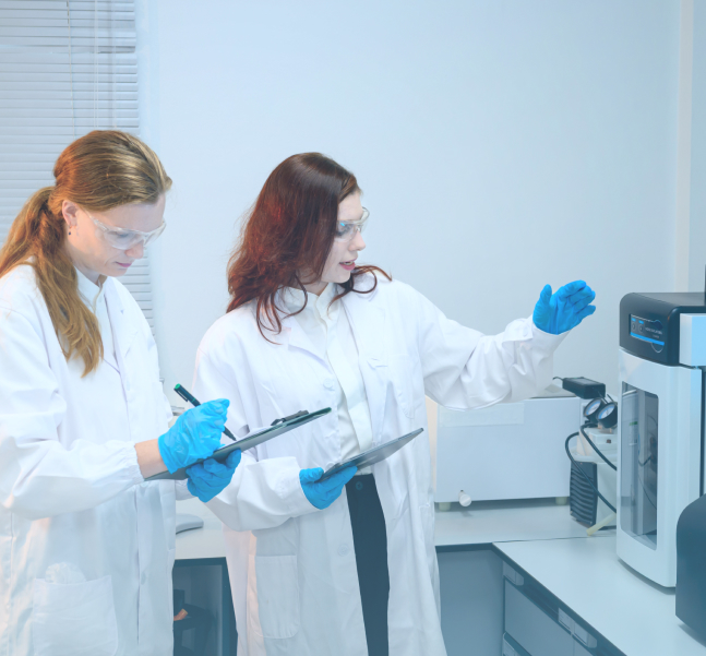 Two female scientists in white lab coats and blue gloves examine instruments beside laboratory equipment, conveying focused collaboration and research.