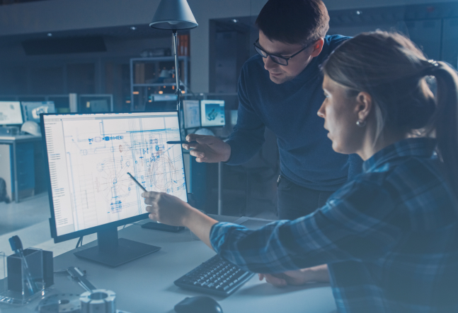 Two engineers review a technical blueprint on a computer monitor, pointing at design details in a dim, focused workspace.
